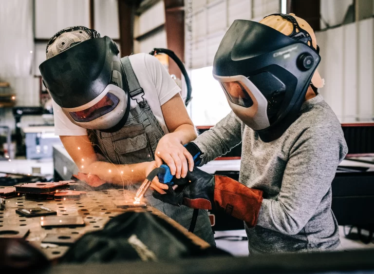 Dos personas con equipo de protección realizan una práctica de soldadura bajo supervisión. La imagen resalta la importancia de la capacitación en el manejo de máquinas de soldar para garantizar la seguridad y precisión en los procesos de unión de metales.